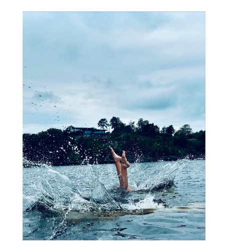 En person står på huvudet i vatten med stänk, omgiven av grönskande landskap.