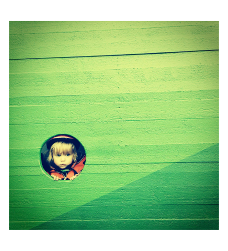 Litet barn i orange jacka kollar fram bakom en grön vägg.